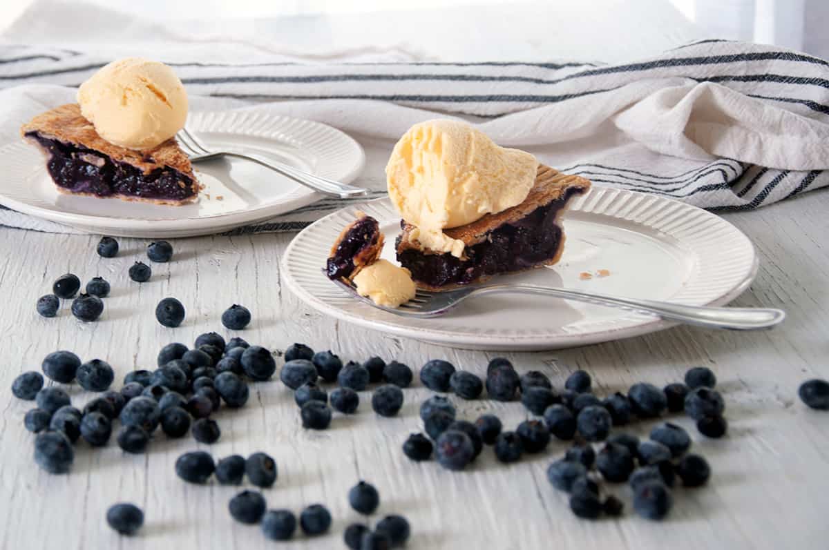 Two slices of blueberry pie on white plates with fresh blueberries all around.