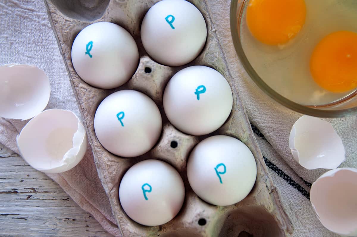 Half a dozen pasteurized eggs in an egg carton with two cracked eggs in the background.