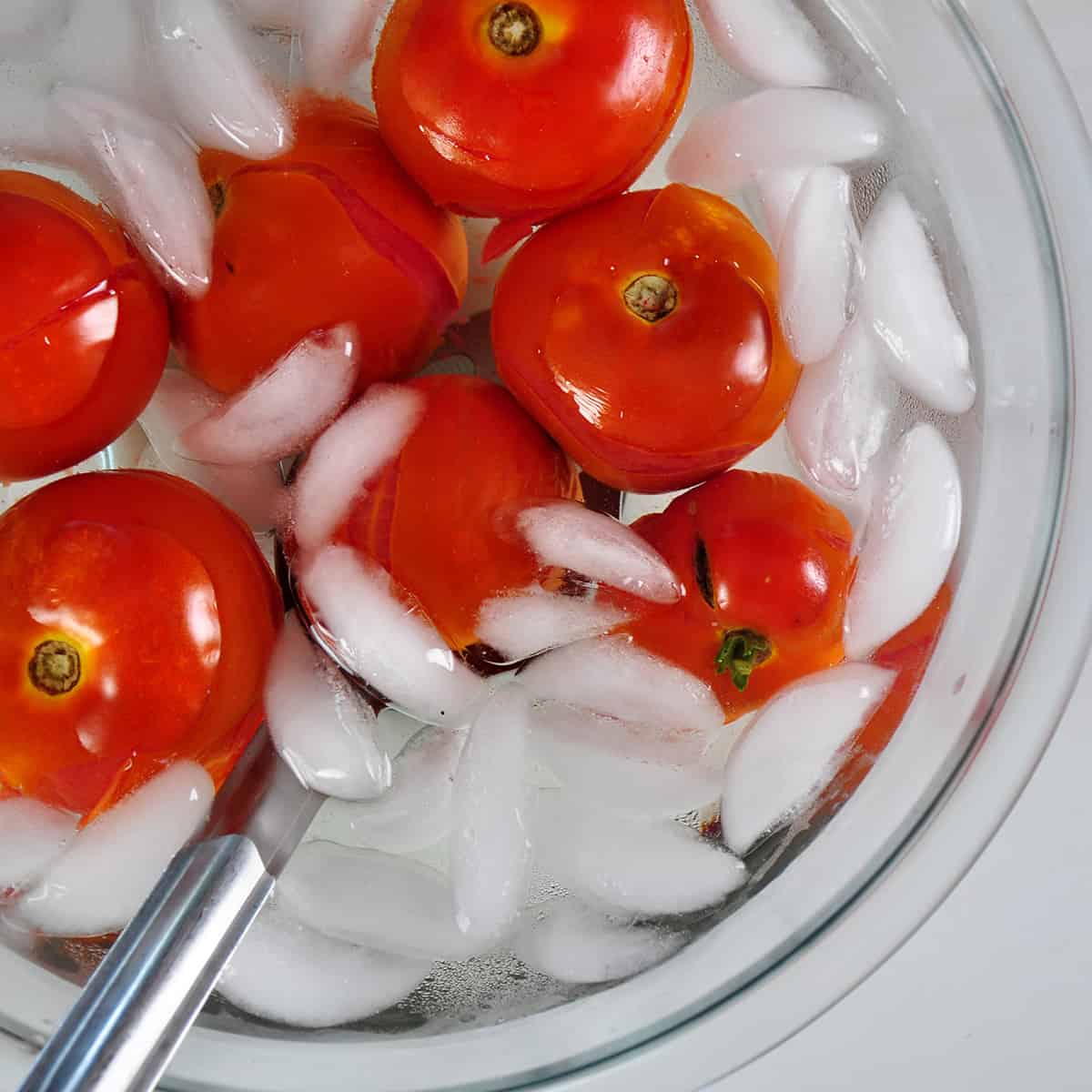 A large glass bow filled with water, ice, and tomatoes whose skin is peeling away.