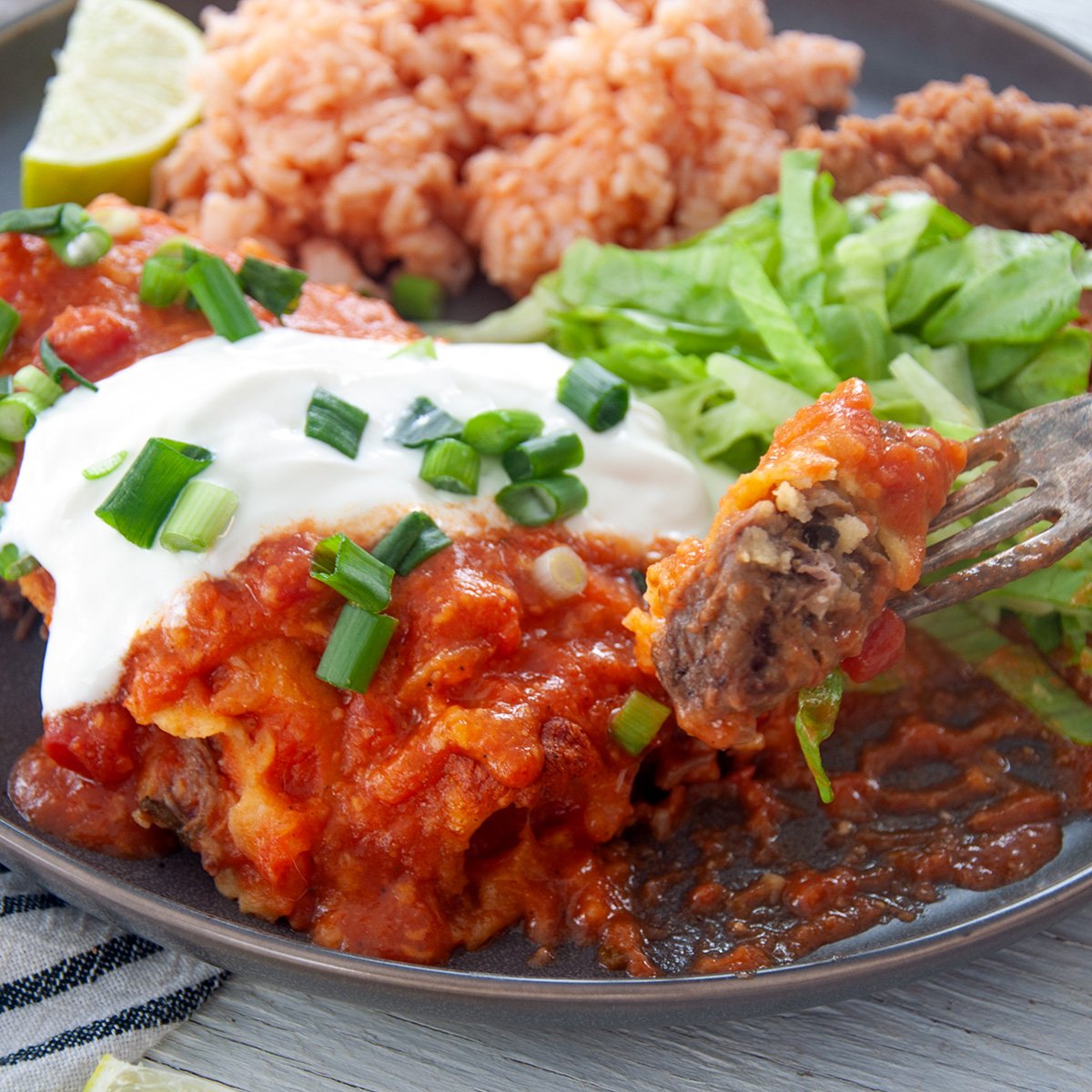 A fork with a single bite of shredded beef enchilada with the whole serving plate behind it.
