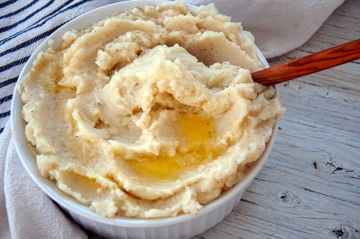 Mashed potatoes in a big container with melted butter and a wooden spoon.