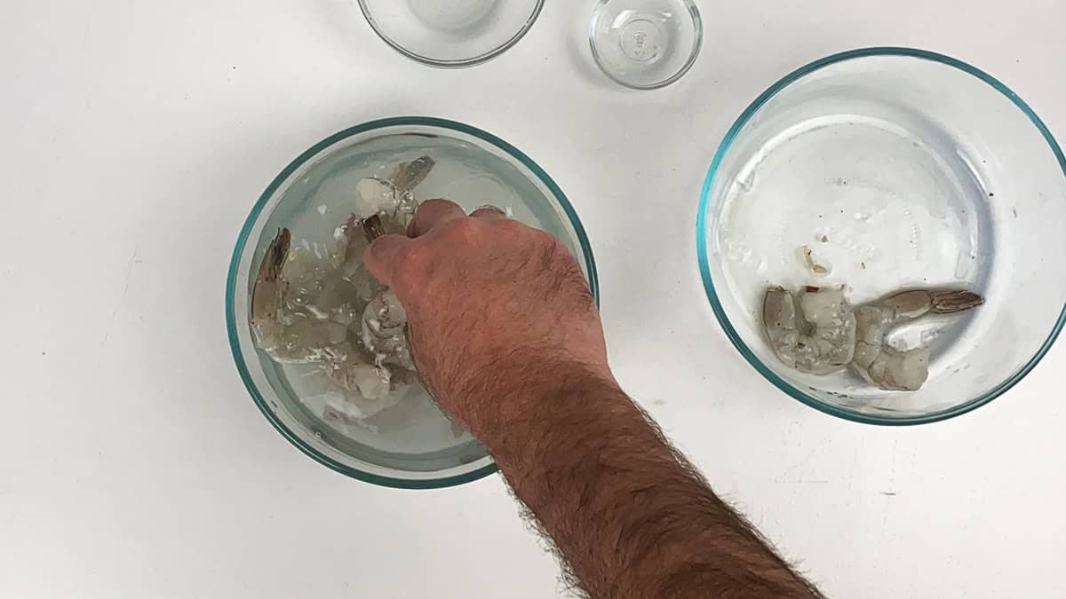 A hand placing raw shrimp into a bowl full of water, sugar, and salt.