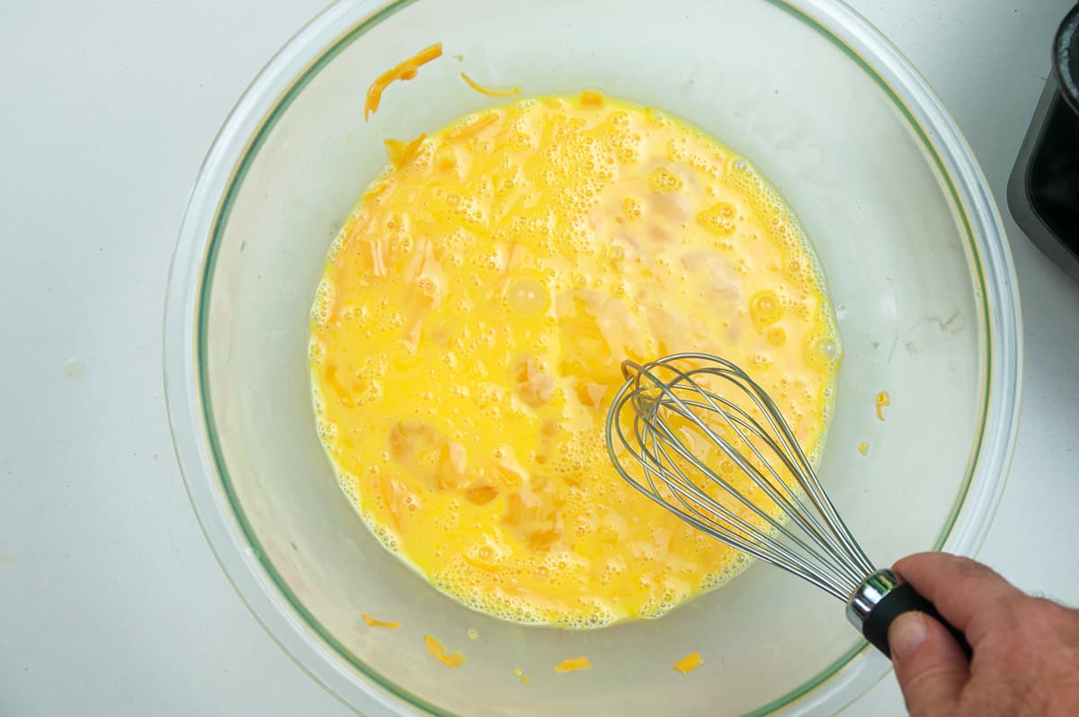 Eggs, milk, and cheese in a large glass bowl with a whisk in it.