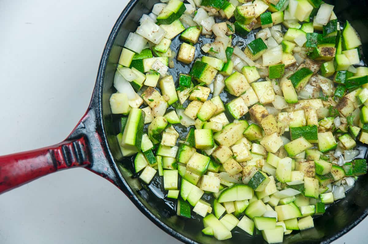 A cast iron containing frying zucchini, onions, and herbs.