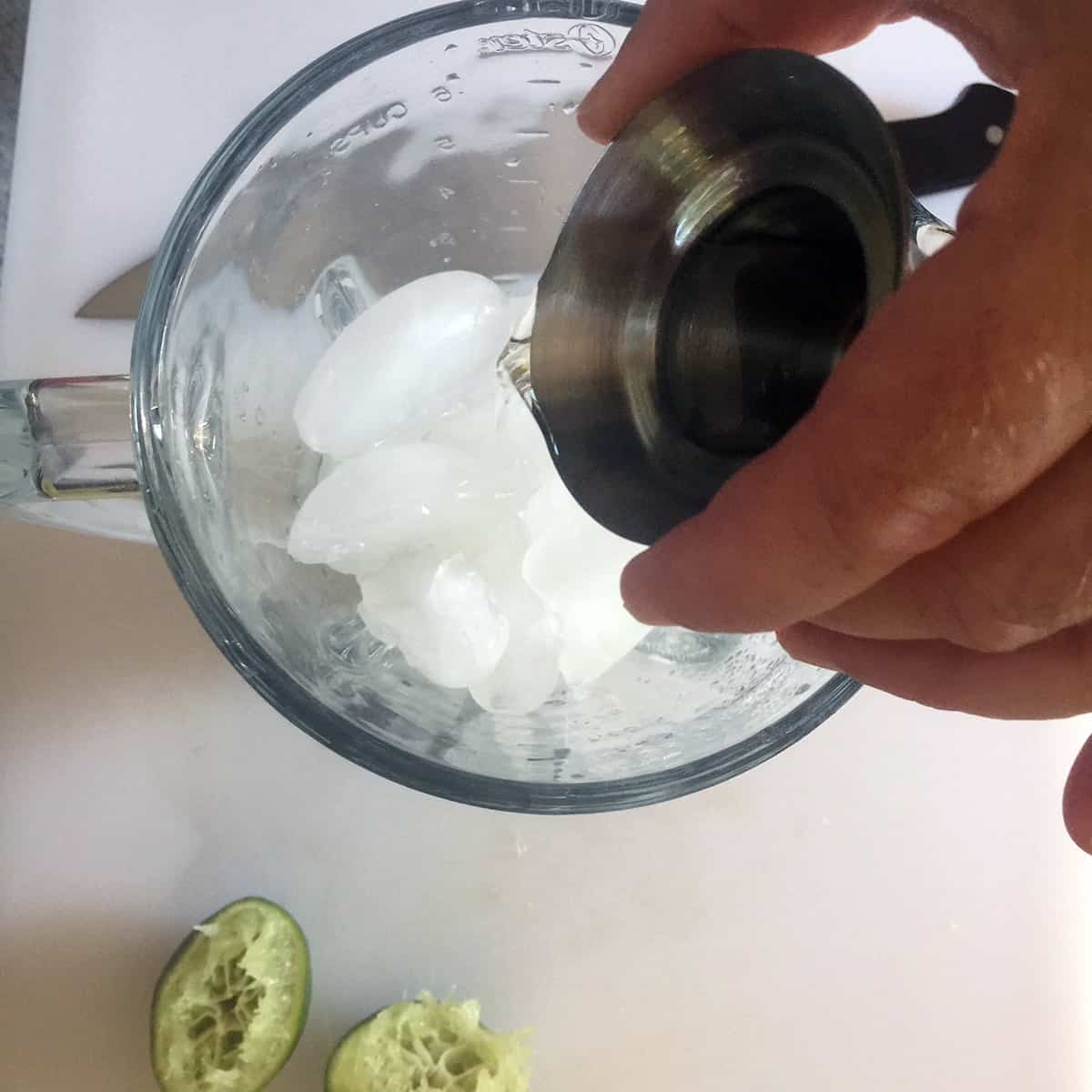 A blender with ice and a hand pouring in a jigger of tequila.