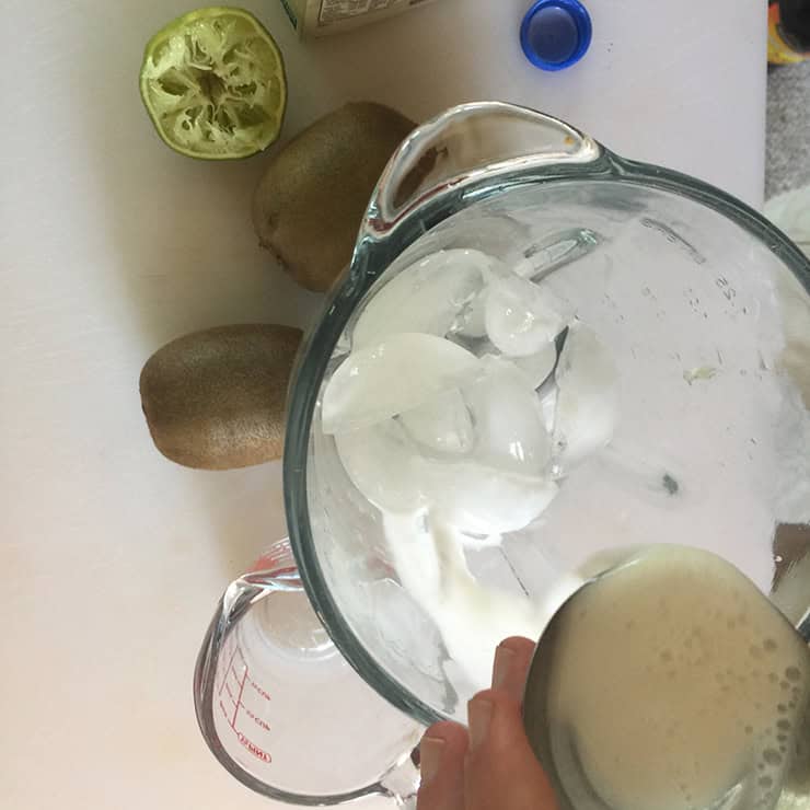 Pouring coconut milk into the blender in preparation for making a margarita. There are two whole kiwis in the background, a spent lime, and a measuring cup.