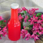 Rhubarb Syrup in a bottle on a picnic table with flowers laying around.