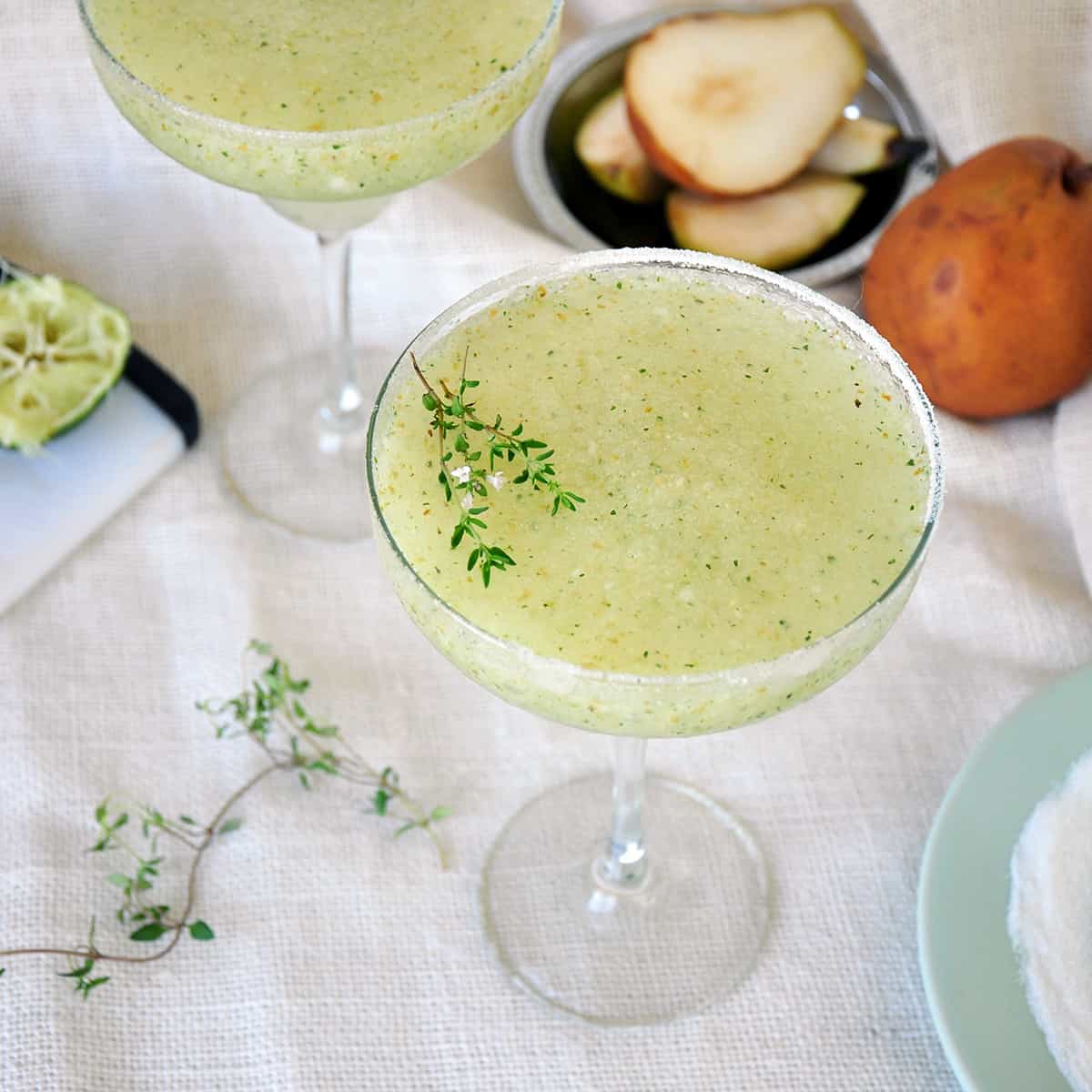 This is a Frozen Pear and Thyme Margarita with thyme, a whole pear, sliced pear, and a spent lime in the background.