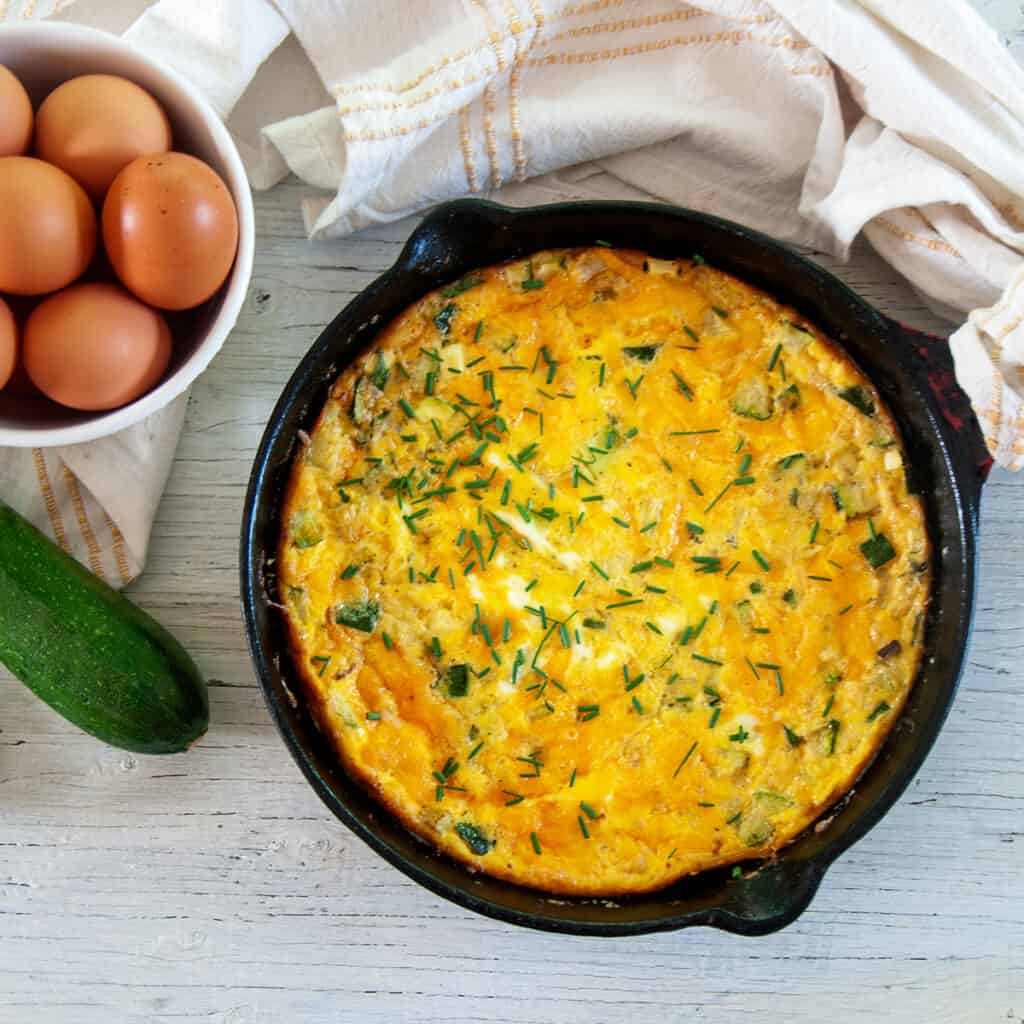 Cast iron skillet with Zucchini Frittata and whole eggs and a raw zucchini to the side.