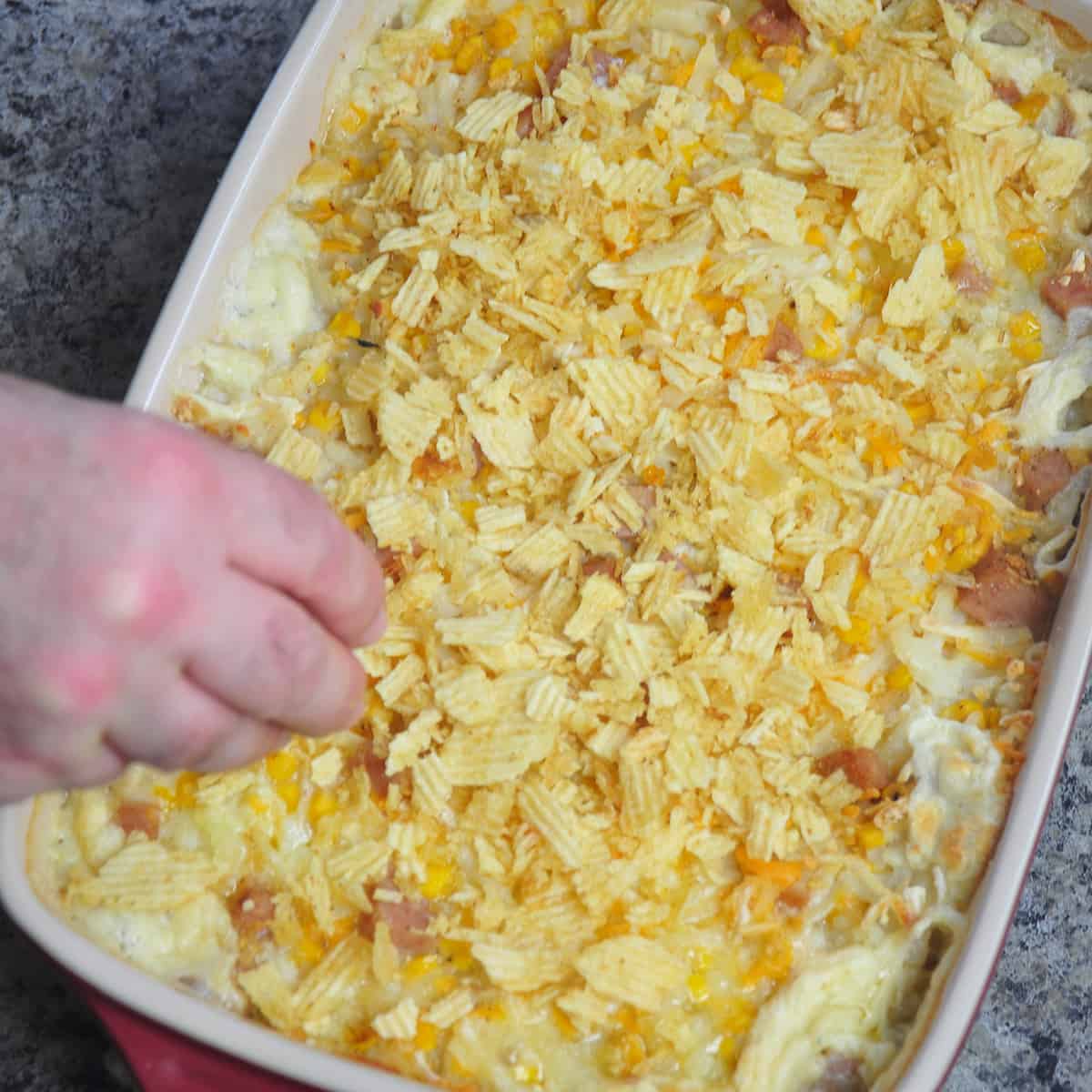 A hand sprinkling crumbled potato chips over casserole.