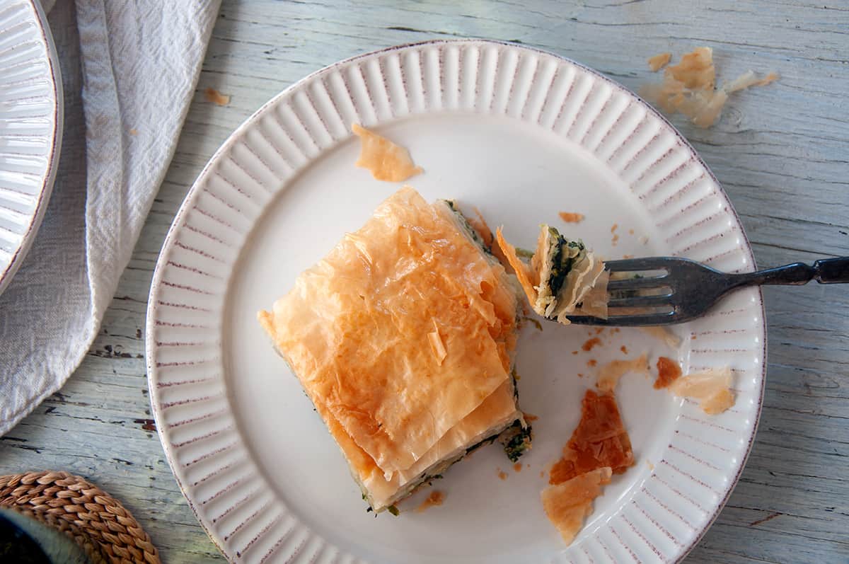 Piece of Spanakopita on a white plate with a fork holding one part.